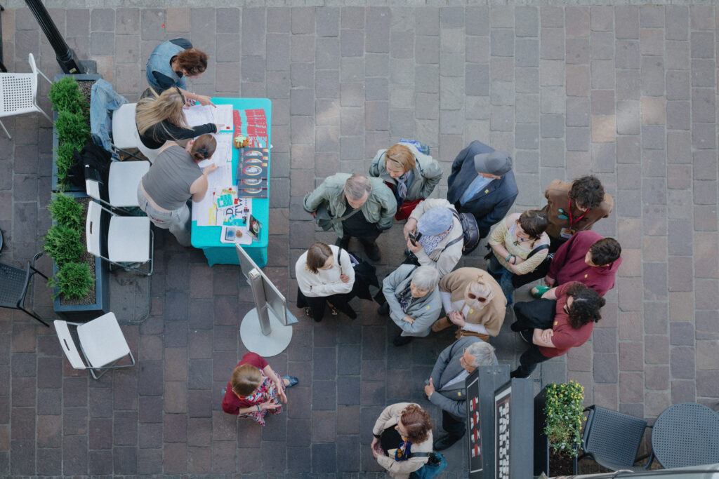 Widok z góry na punkt informacyjny Dni Dziedzictwa. Wolontariusze pochylają się nad stolikiem, a obok zebrała się grupka uczestników.