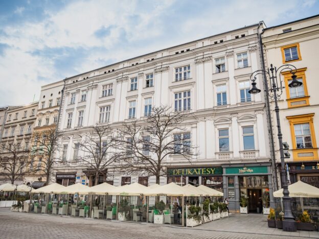 Ulica miejska z rzędem zabytkowych kamienic, przed którymi stoją ogródki kawiarniane pod jasnymi parasolami, bezlistne drzewa oraz duża latarnia uliczna, a w tle widać częściowo zachmurzone niebo i przechodzących ludzi.