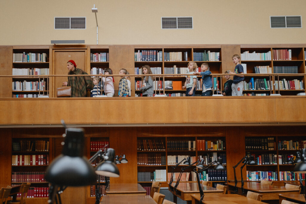 W bibliotece pełnej drewnianych półek z książkami dorosły mężczyzna z dziećmi stoi na górnej galerii, obserwując poniżej rzędy stołów do czytania z lampkami.