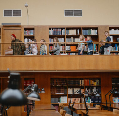 W przestronnej bibliotece z drewnianymi regałami pełnymi książek, na górnym balkonie stoi dorosły z grupą dzieci, podczas gdy na niższym poziomie widać stoły do nauki z lampami.